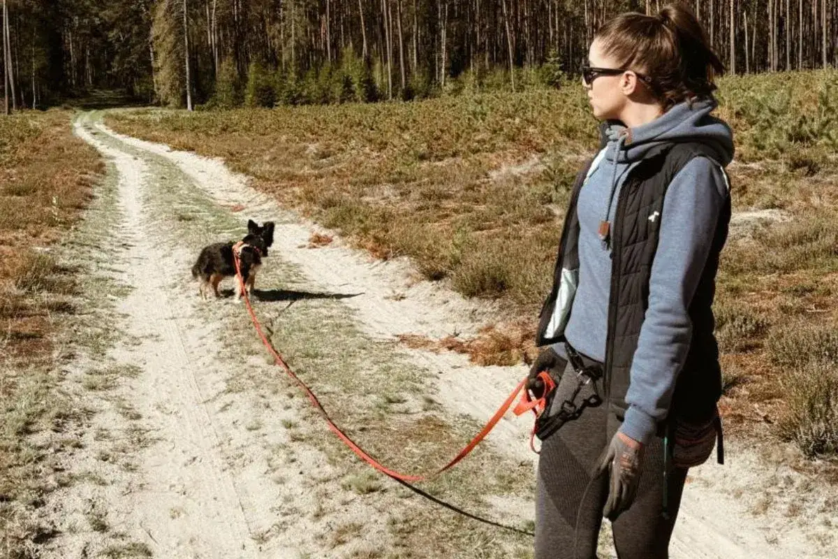 Kobieta z psem na smyczy na leśnej ścieżce. Wygodny str&oacute;j i saszetka to podstawa, gdy zastanawiasz się, co wziąć na spacer z psem.